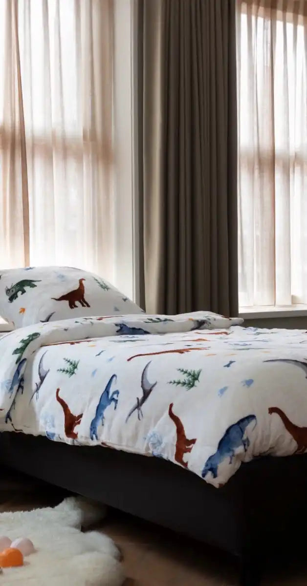 Een bed met een kleurrijk dekbedovertrek met dino's in een sfeervolle kamer, bekleed met zachte velvet stof. Het dino-patroon springt eruit tegen de luxe, fluweelachtige achtergrond van de kamer. foto: dekbedovertrek dino collections.