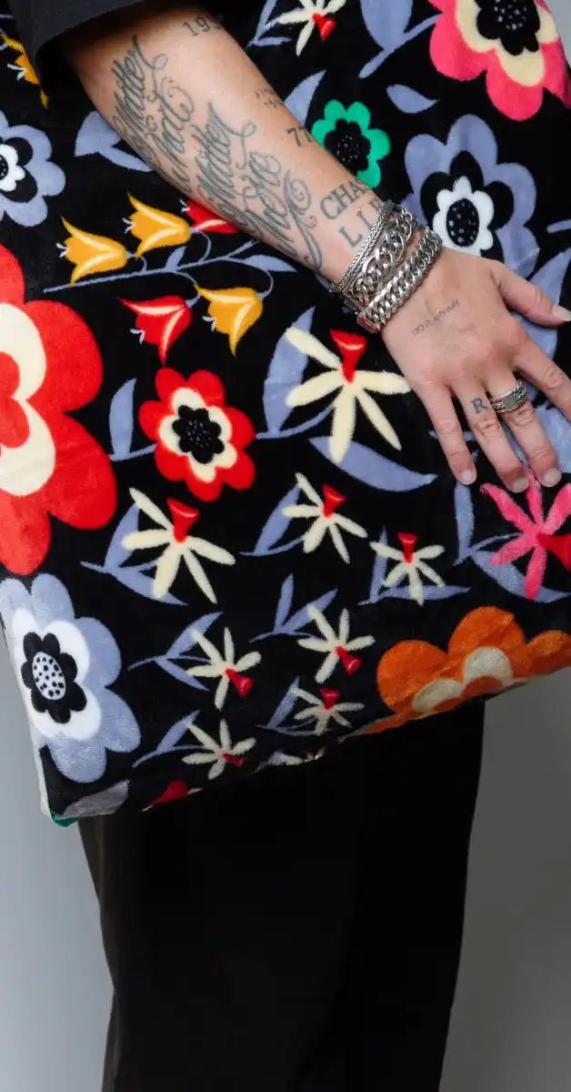 Close-up van een hand op een kussensloop met bloemenpatroon van zachte velvet stof. Het ontwerp toont kleurrijke bloemen in rood, oranje, geel, groen en paars op een zwarte achtergrond. foto: kussensloop bloemen collecties.