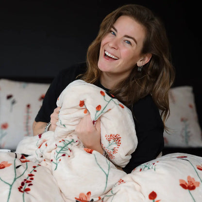 Een vrouw zit ontspannen met een velvet tweepersoons dekbedovertrek om haar heen gedrapeerd. Het dekbedovertrek heeft een kleurrijk patroon van levensechte klaprozen en straalt luxe uit. foto: dekbedovertrek klaproos met Lisa.