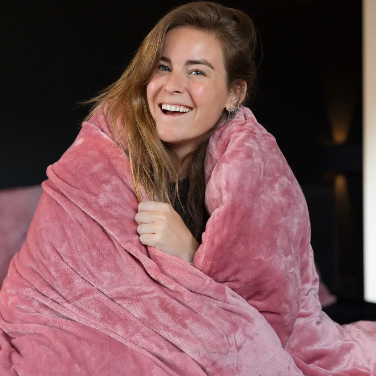 Een vrouw gewikkeld in een zacht, roze velvet dekbedovertrek. Het materiaal glanst subtiel in het licht, terwijl ze zich knus en warm voelt, omgeven door de luxe en zachtheid van de fluweelachtige stof. foto: dekbedovertrek roze met Lisa.