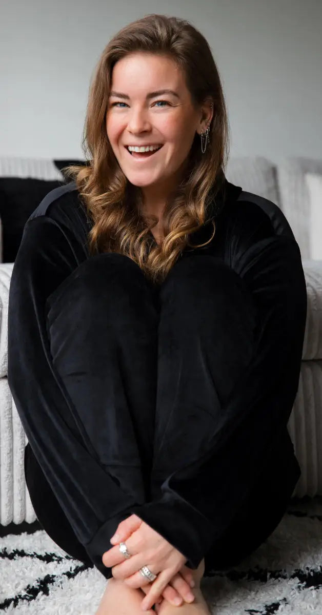 Een vrolijke meid draagt een huispak  zwart velvet huispak ze heeft een ontspannen houding terwijl ze op de grond zit, met haar handen om haar voeten geslagen.
foto: huispak zwart met vrouw